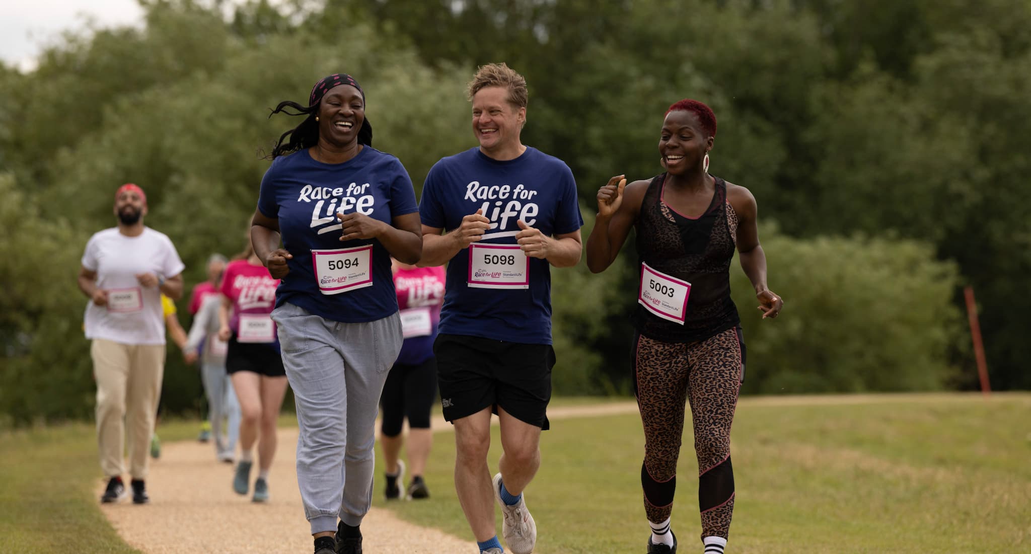 A man is running in park and wearing a blue T-shirt, while taking part in a Race for Life 10K event. He looks happy.