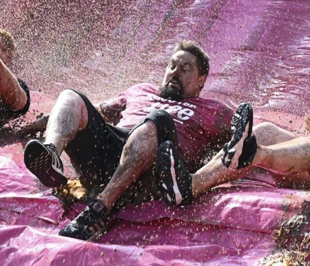 two men and a woman slide down a pink slide into muddy water making a splash of mud.